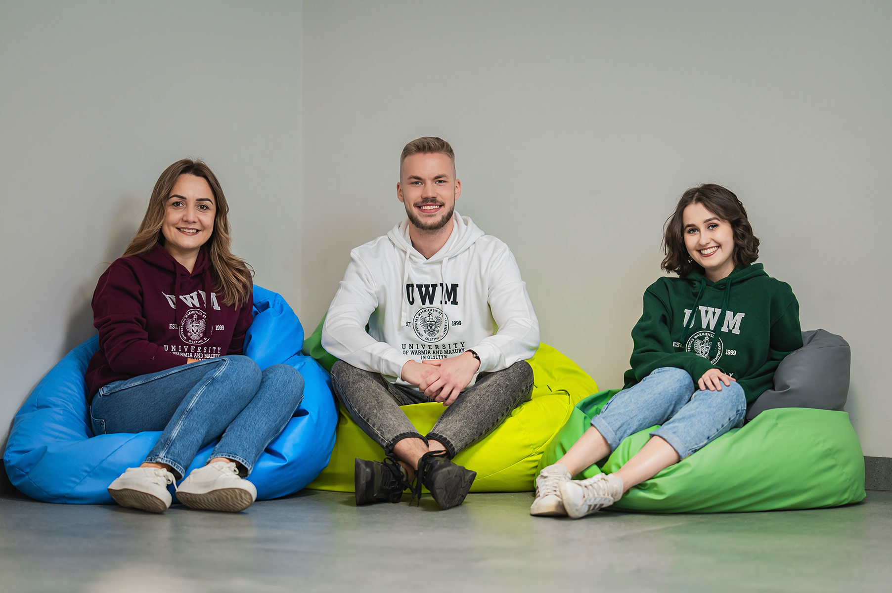 Trzy osoby siedzą na pufach w pomieszczeniu i uśmiechają się do aparatu