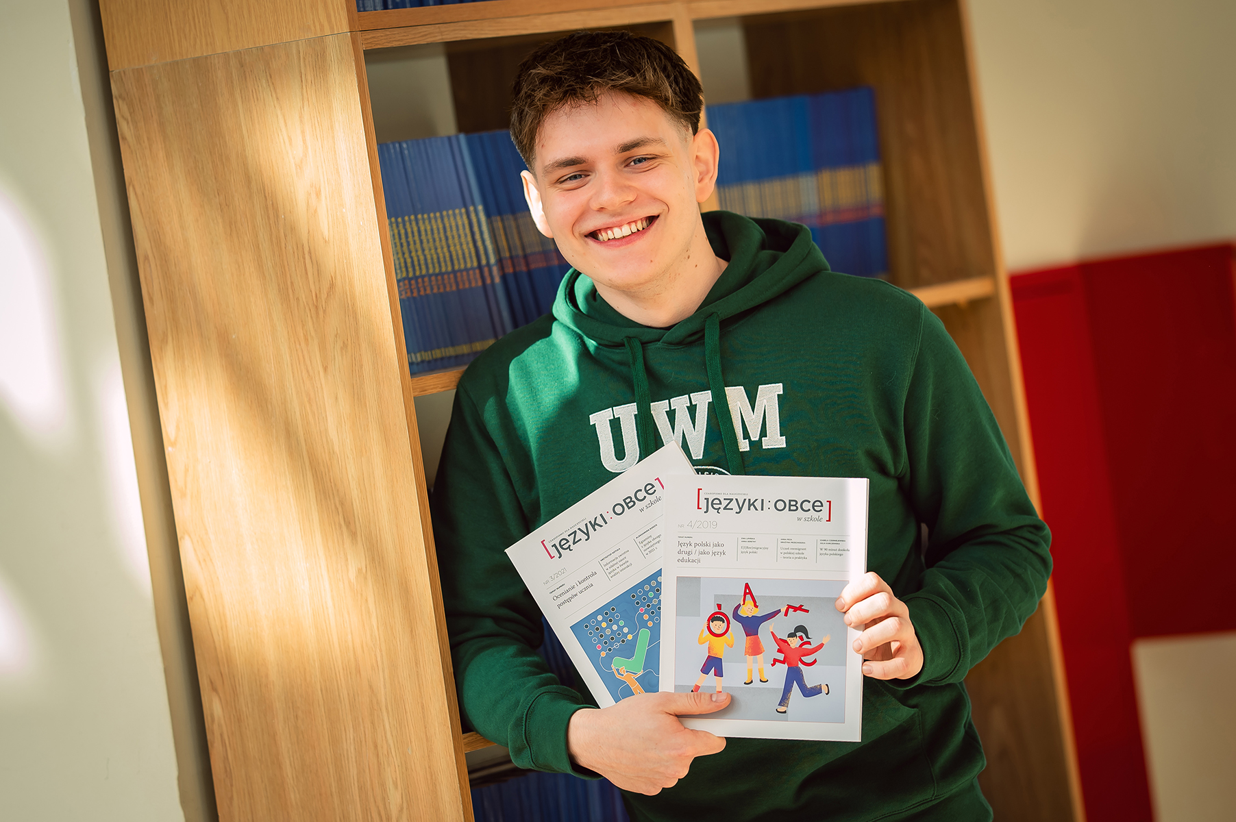 Uśmiechnięty student w zielonej bluzie UWM trzymający dwa numery czasopisma „Języki Obce”