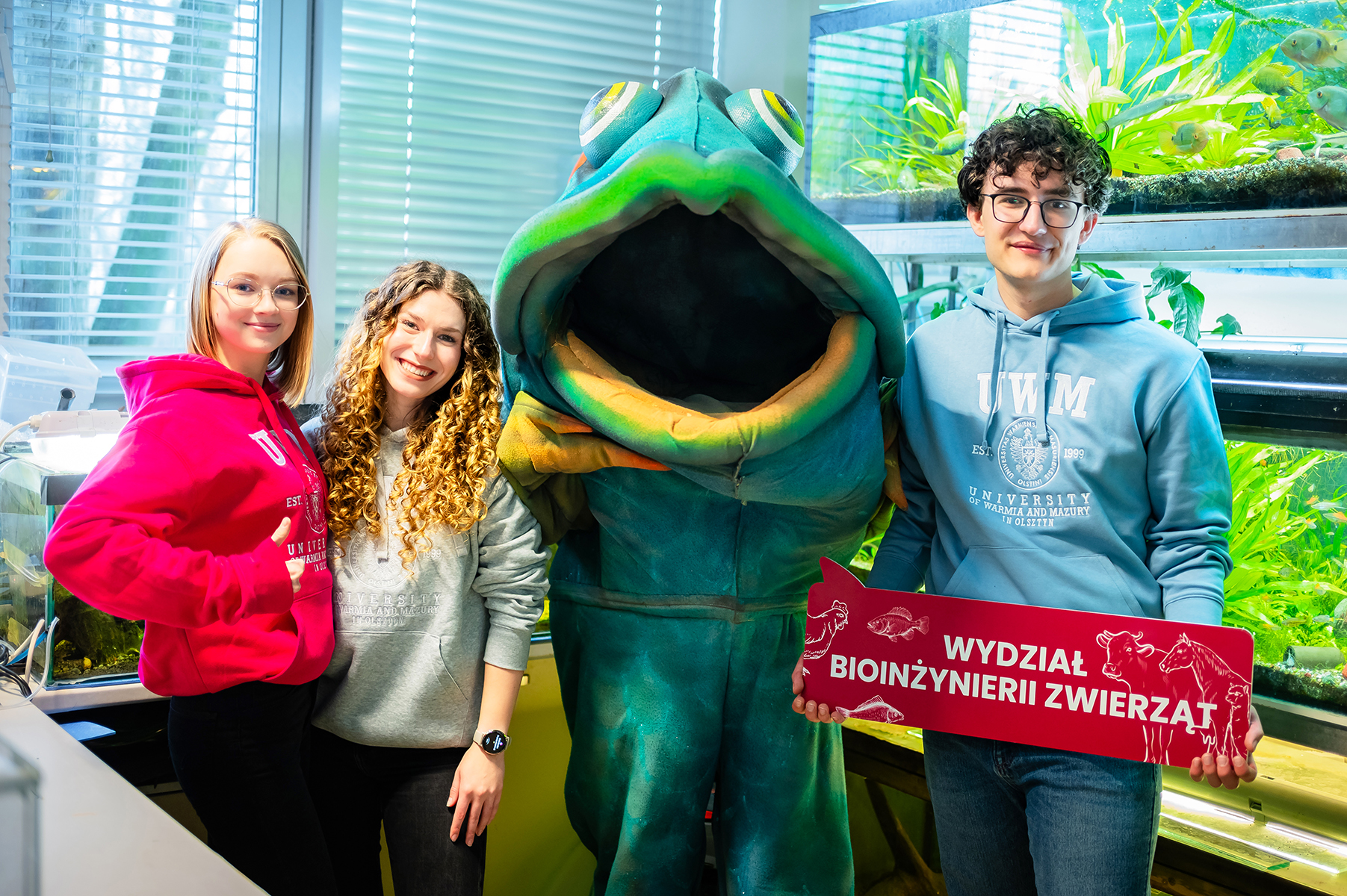 Studenci z maskotką ryby i tablicą Wydziału Bioinżynierii Zwierząt na tle akwariów