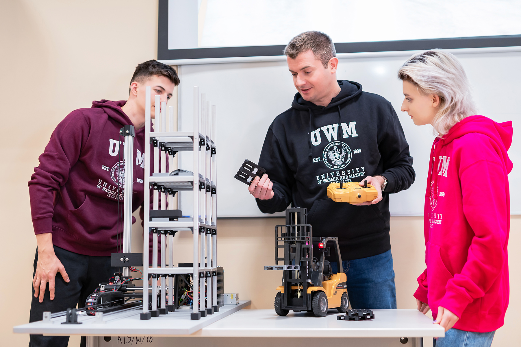 Trzy osoby stoją przy stole i omawiają model wózka widłowego i konstrukcję techniczną