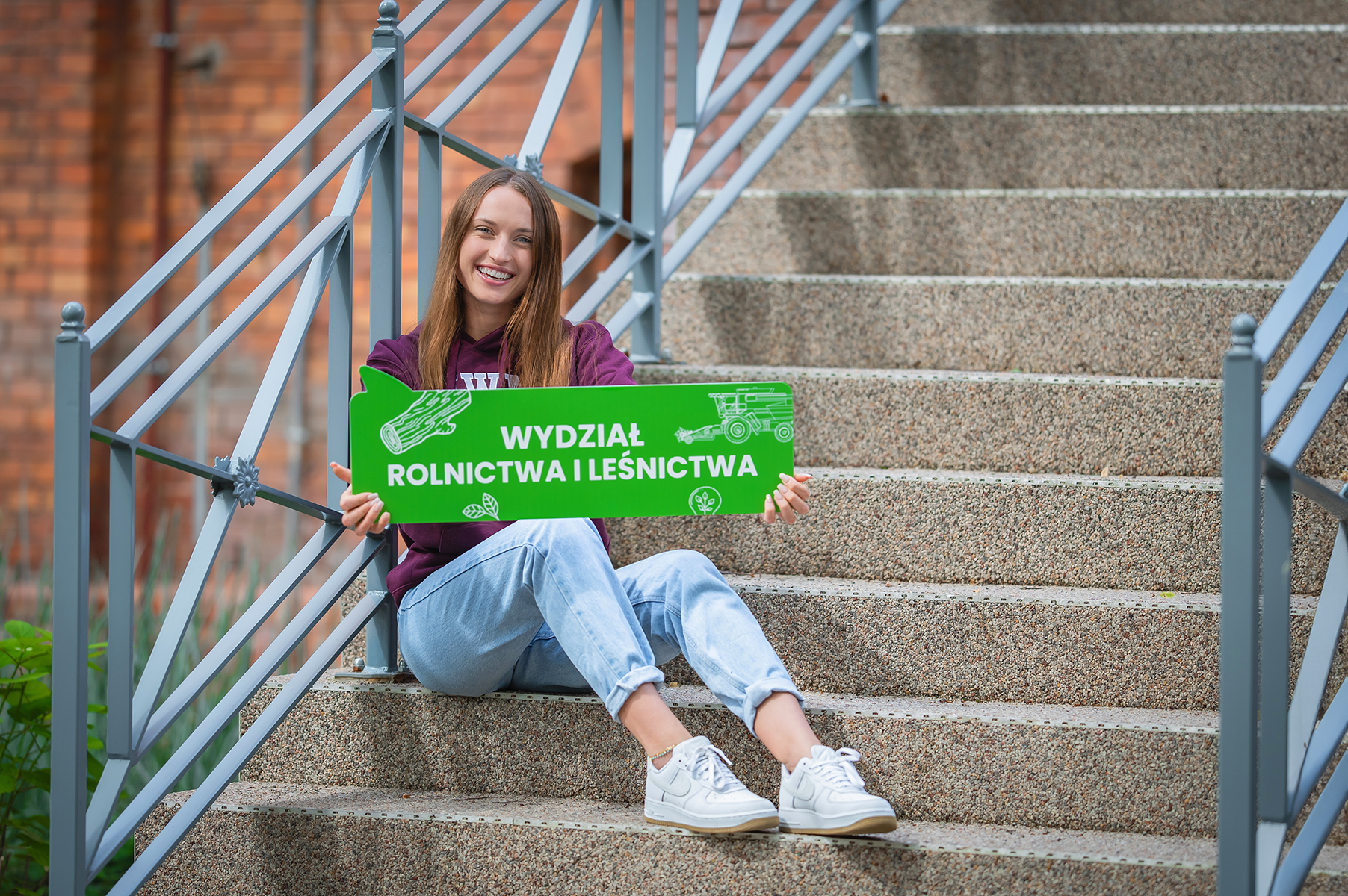 Osoba siedzi na schodach przed i trzyma zieloną tablicę z napisem "Wydział Rolnictwa i Leśnictwa"