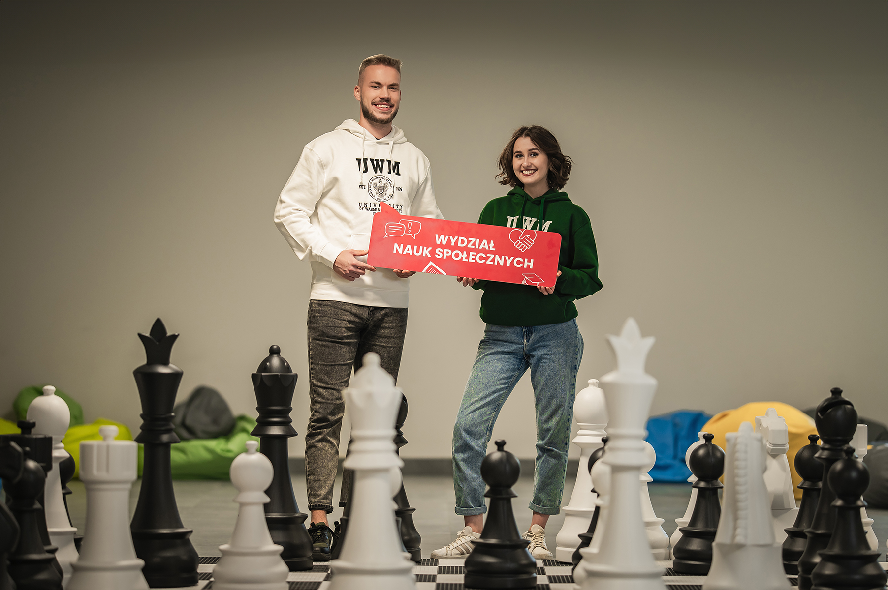 Dwie osoby stoją na dużej szachownicy i trzymają tablicę z napisem „Wydział Nauk Społecznych”