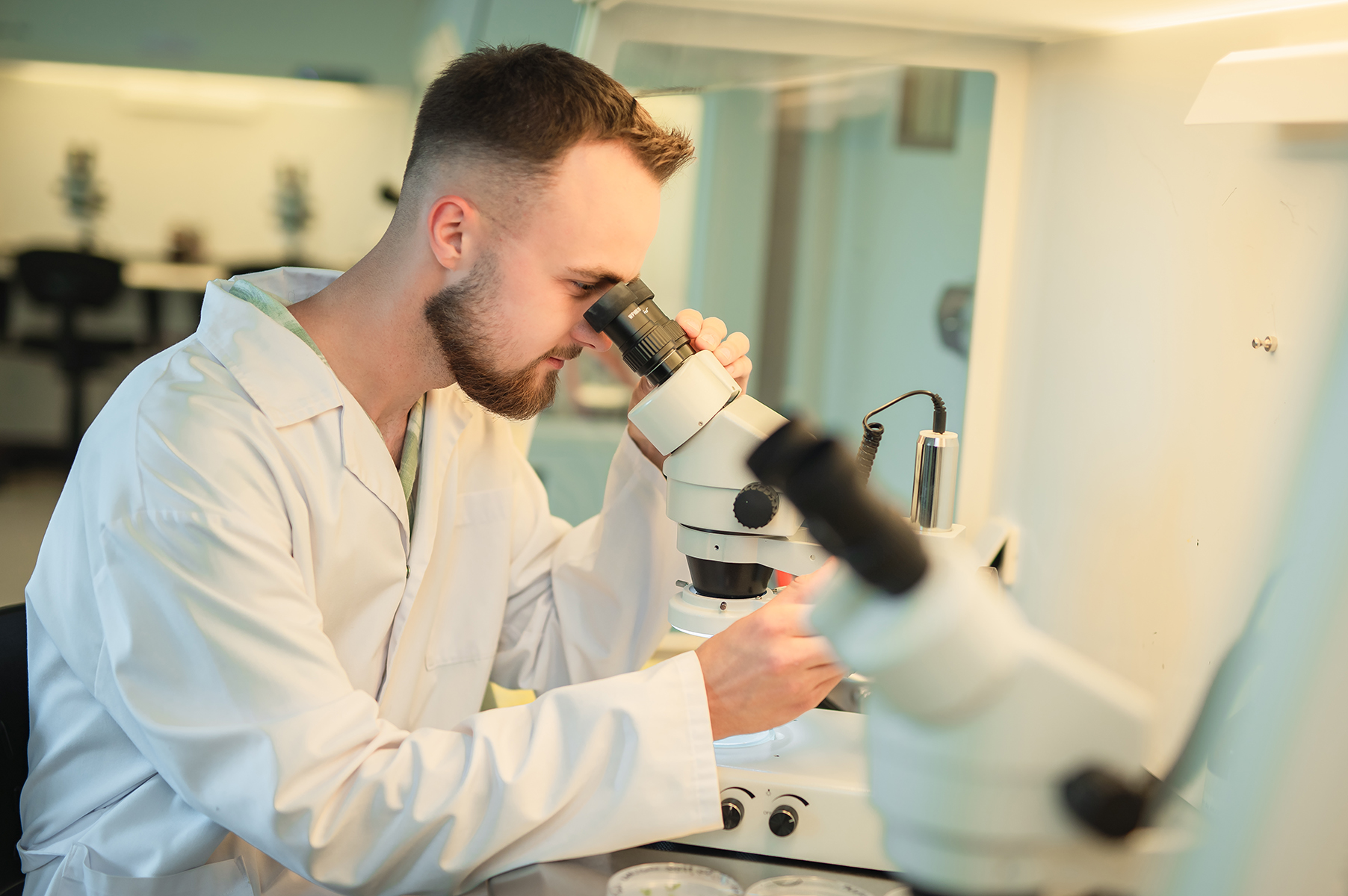 Osoba w fartuchu laboratoryjnym obserwuje próbkę przez mikroskop w laboratorium