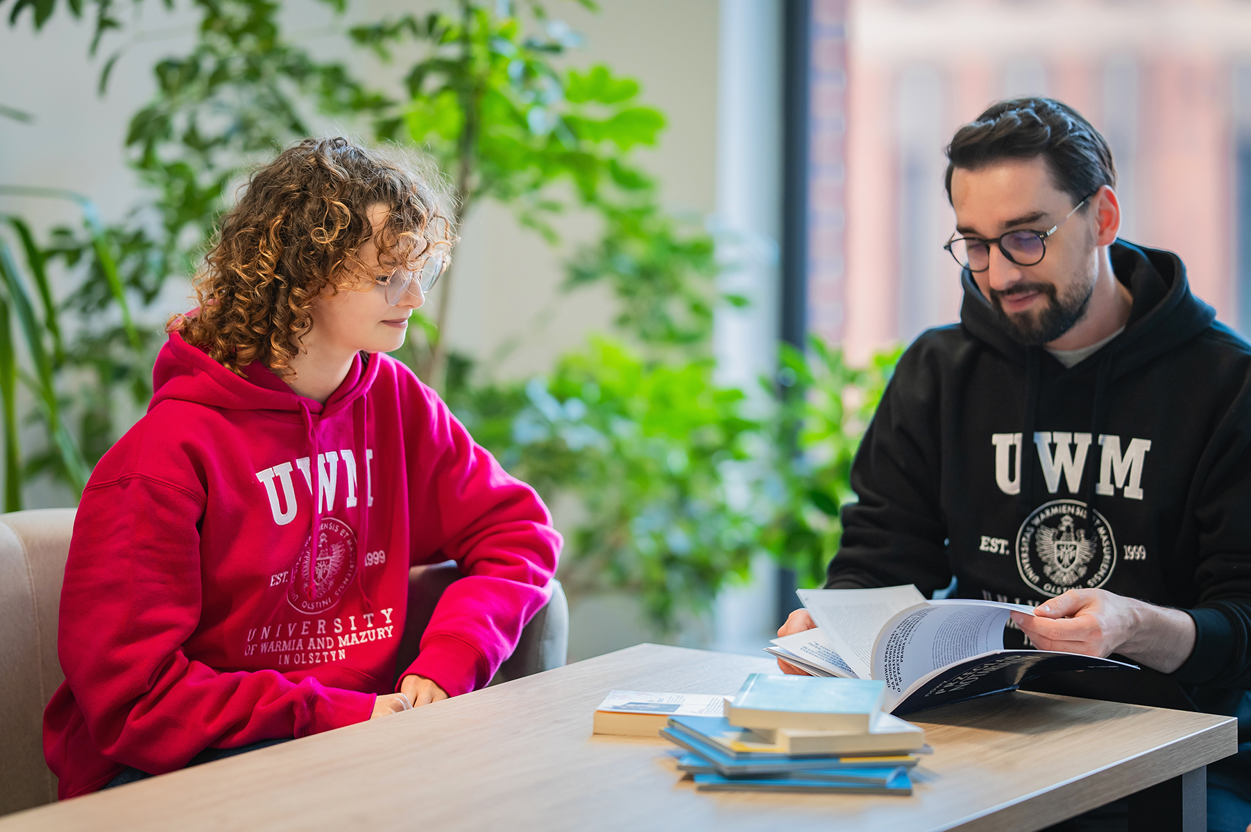 Dwie osoby w bluzach z logo UWM siedzą przy stole i przeglądają książkę
