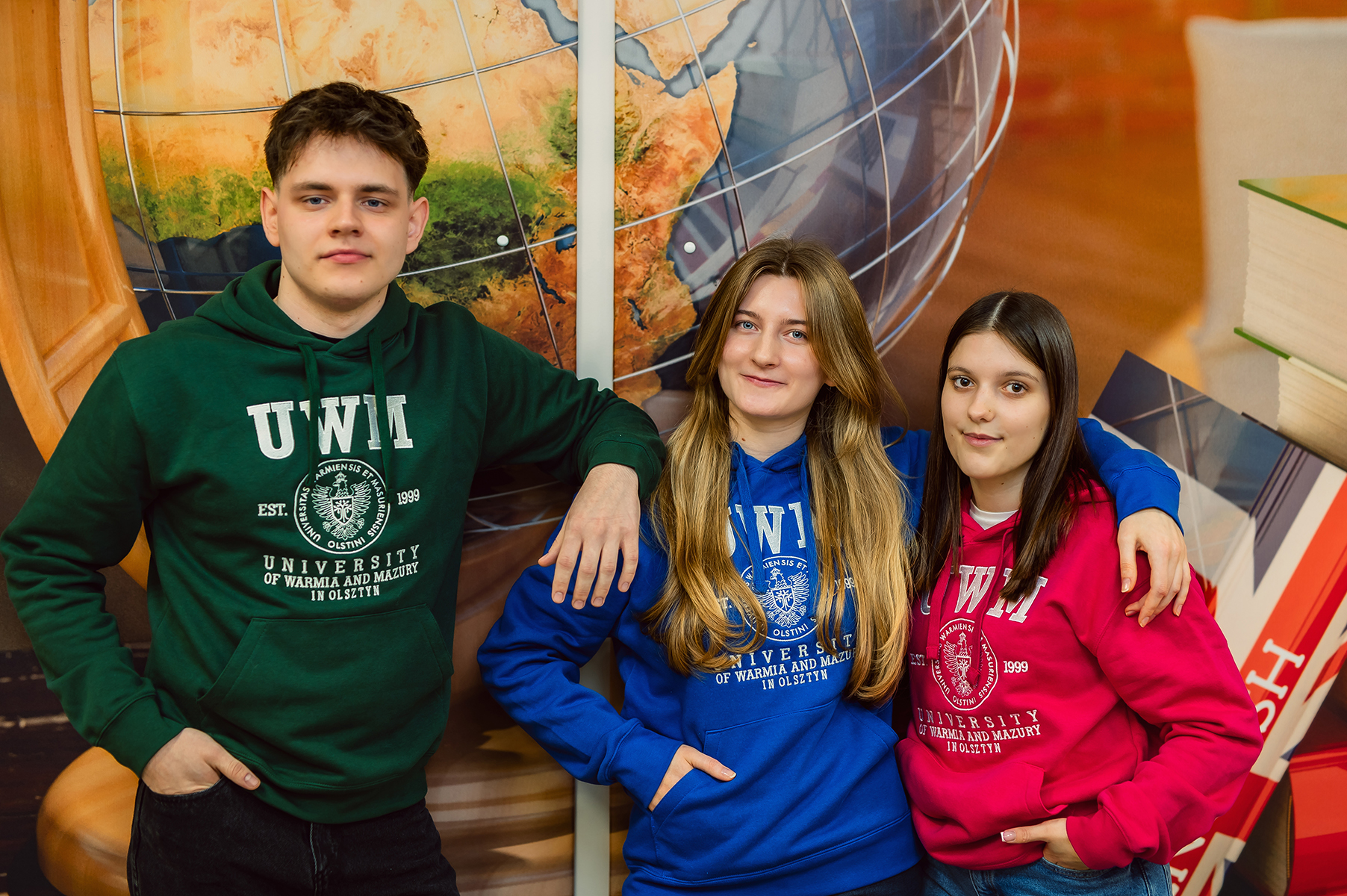 Troje studentów w bluzach uczelni stoi razem na tle grafiki z globusem i książkami