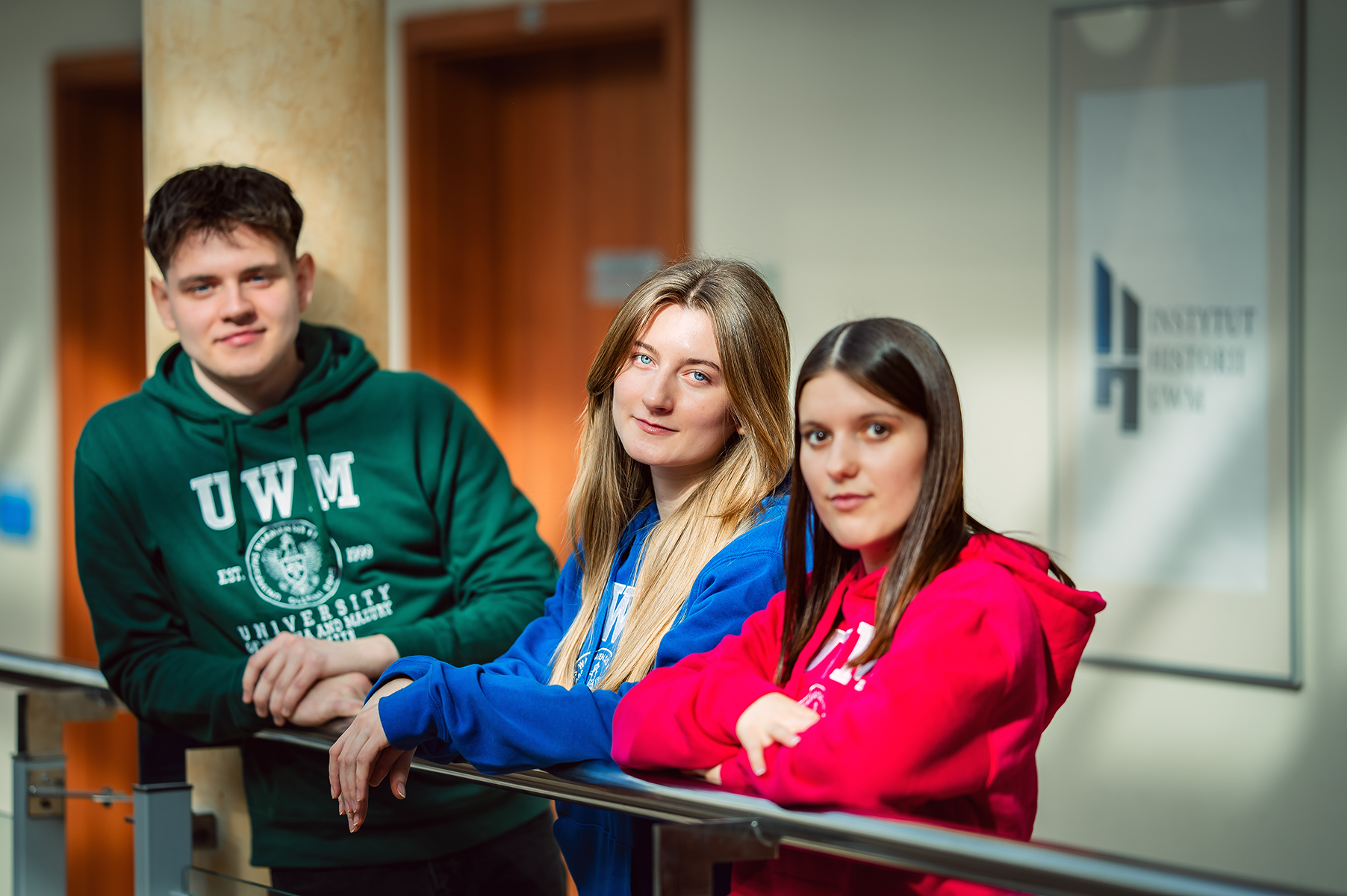 Troje studentów w kolorowych bluzach UWM pozuje na tle szyldu Instytutu Historii UWM
