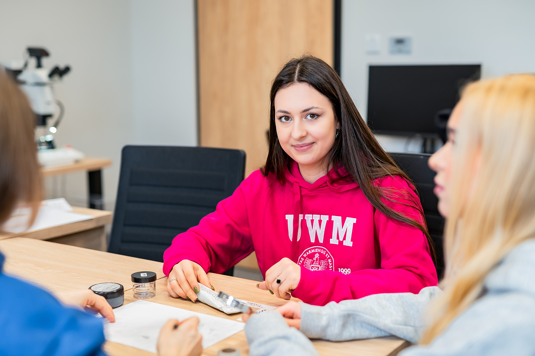 Studentka w różowej bluzie z logo UWM siedzi przy stole i rozmawia z innymi osobami podczas zajęć