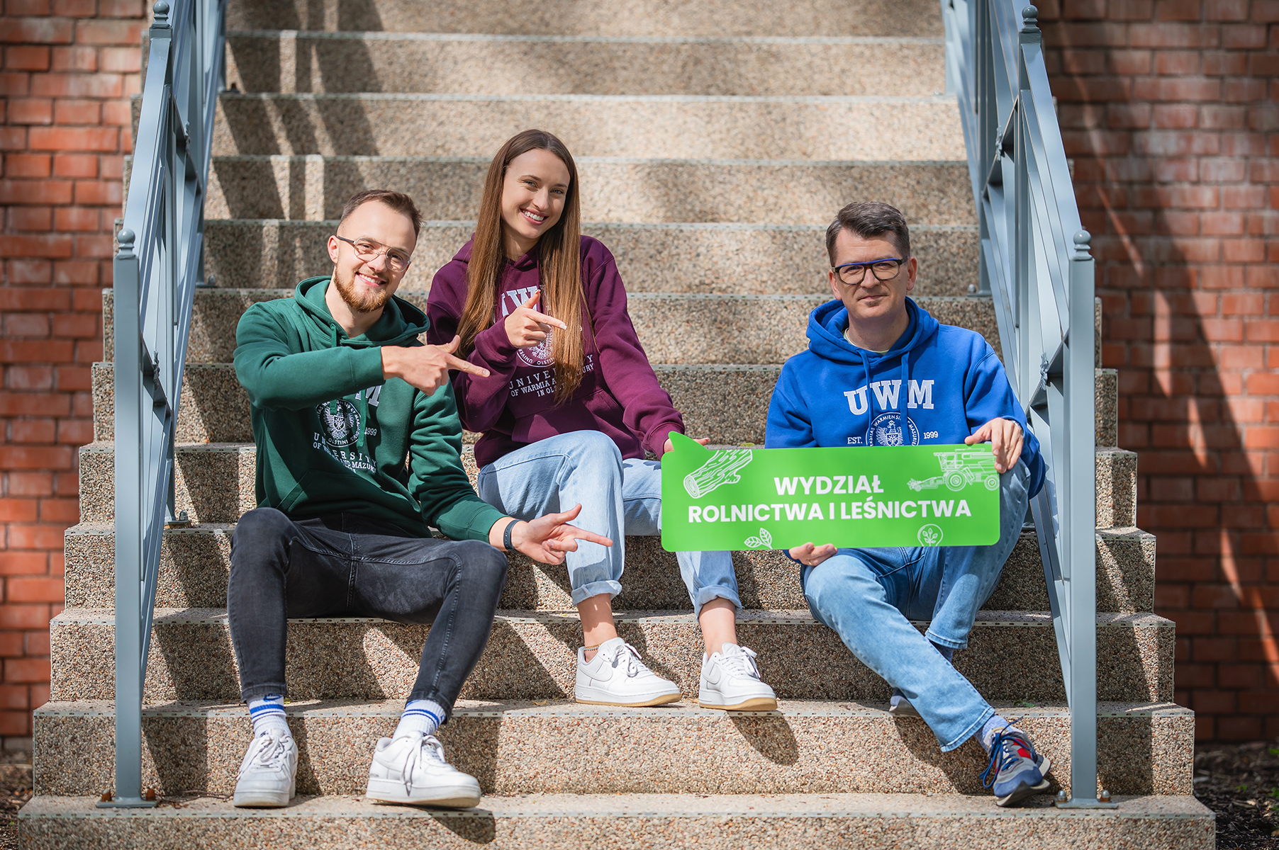 Trzy osoby siedzą na schodach i trzymają tablicę z napisem „Wydział Rolnictwa i Leśnictwa”