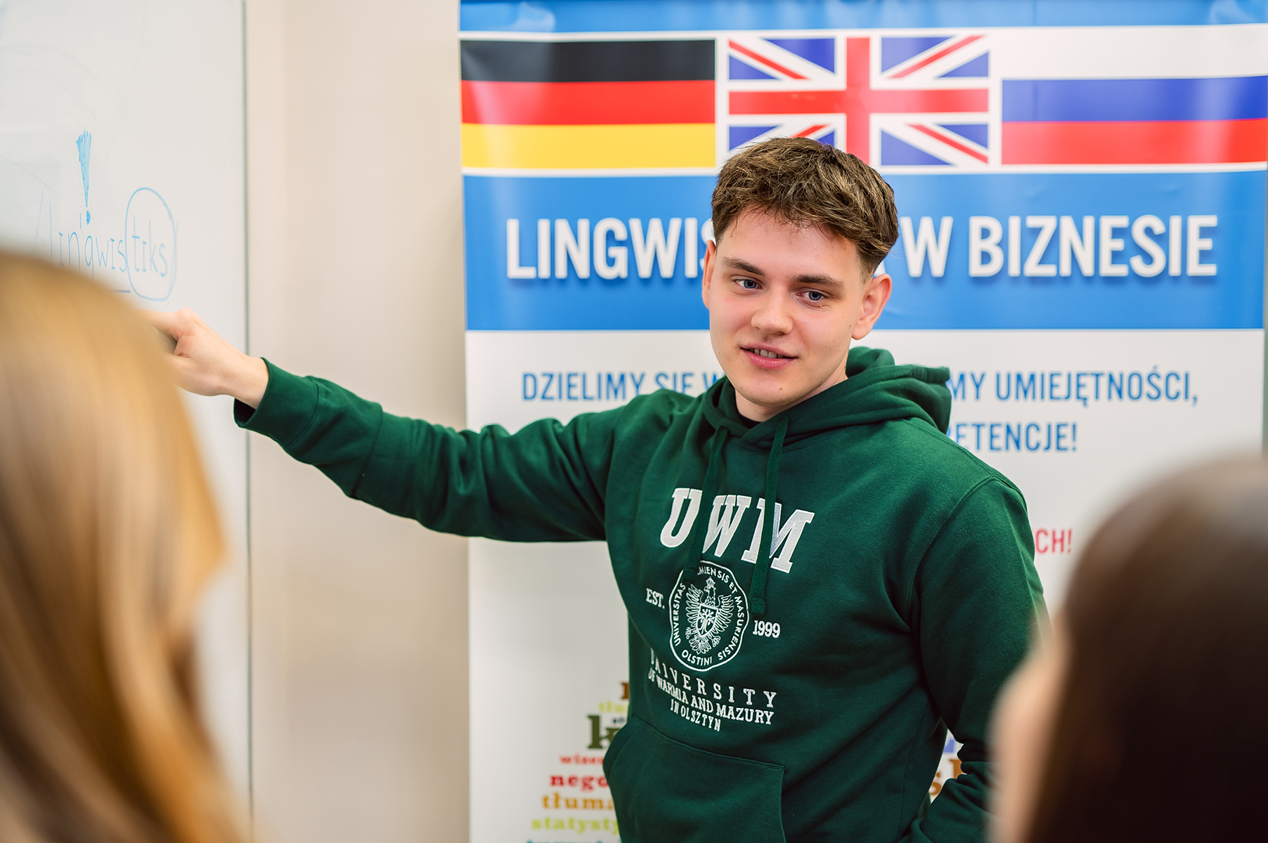 Student w bluzie UWM wskazujący na tablicę obok baneru „Lingwistyka w biznesie”