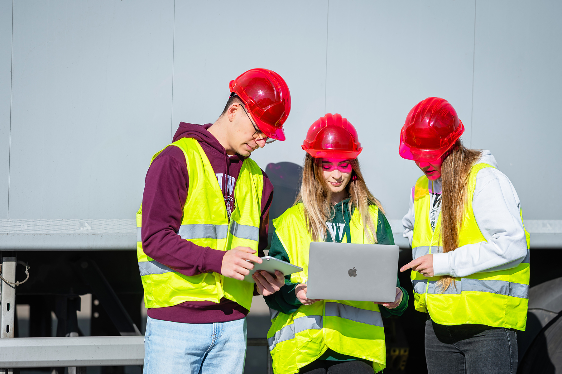 Trzy osoby w kaskach i kamizelkach odblaskowych sprawdzają dane na laptopie przy ciężarówce