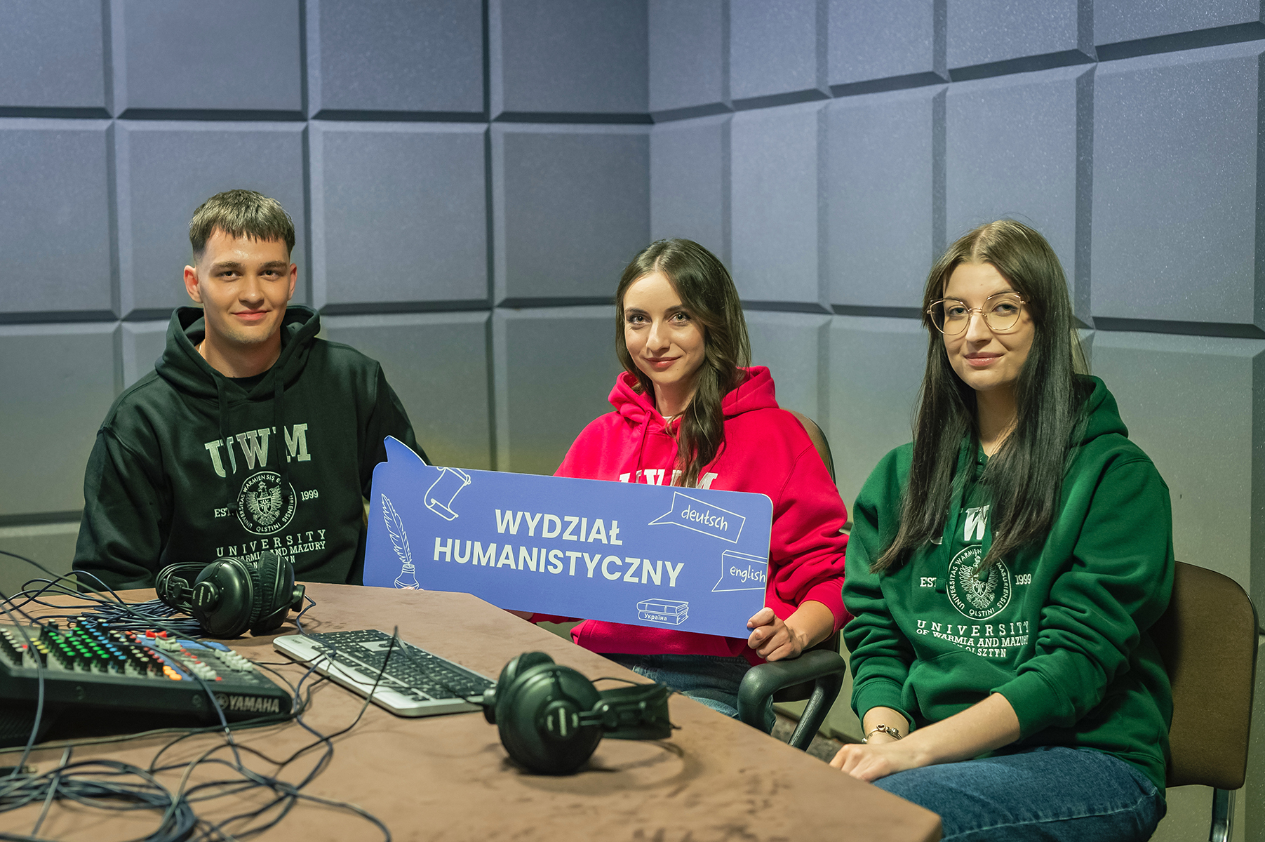 Troje studentów w studiu nagrań trzyma tabliczkę z napisem „Wydział Humanistyczny”