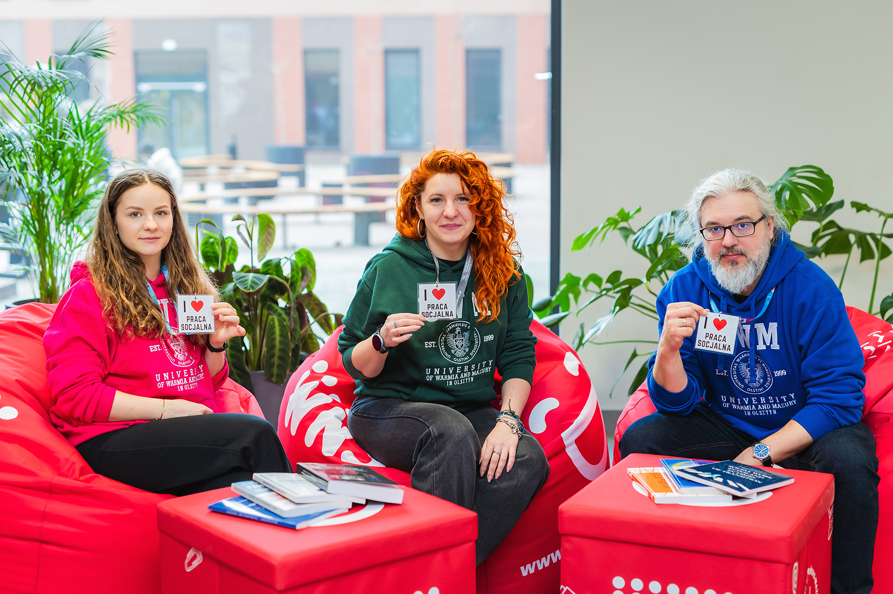 Trzy osoby siedzą na różowych pufach, trzymają kartki z napisem „I love praca socjalna”