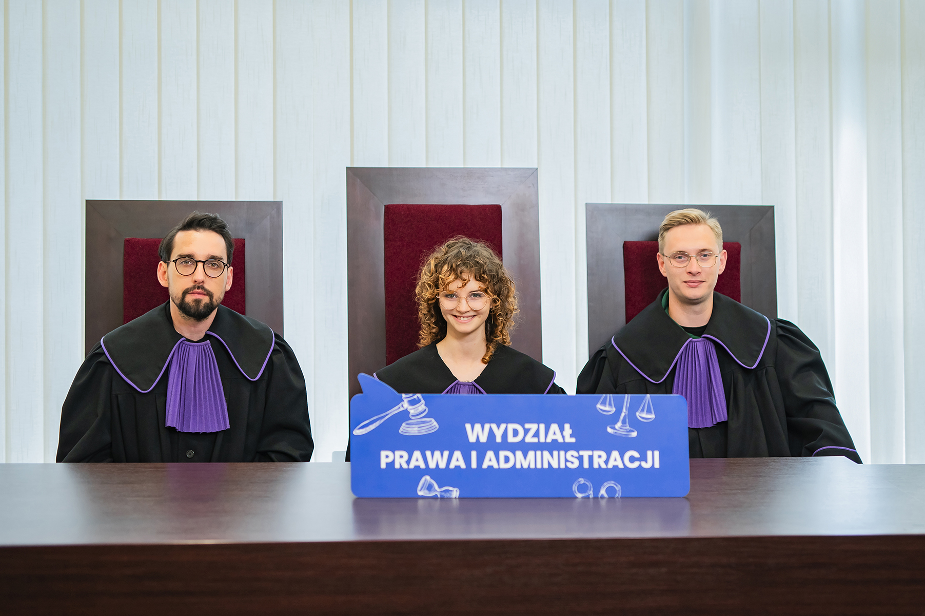 Trzy osoby w togach siedzą za stołem i trzymają tablicę z napisem "Wydział Prawa i Administracji"