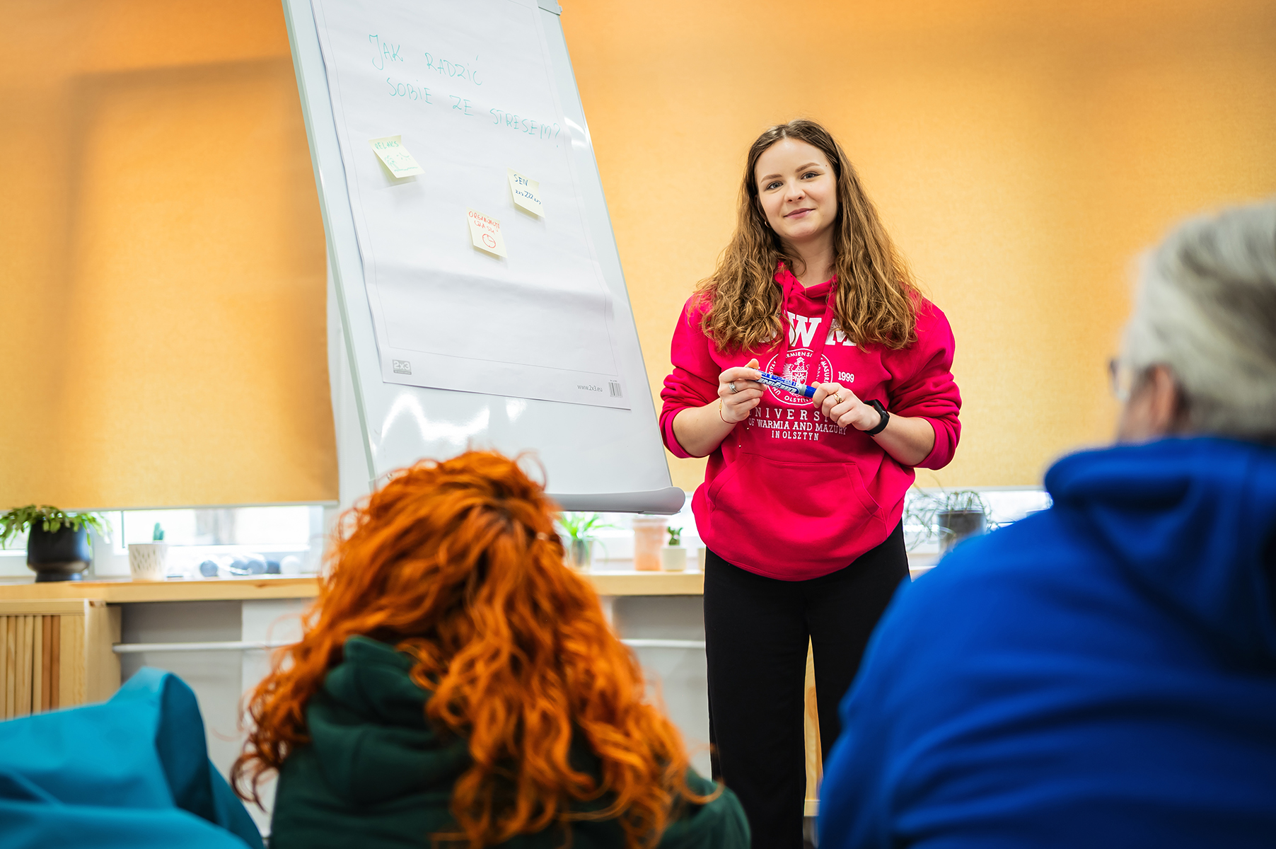 Osoba prowadząca zajęcia stoi przy flipcharcie i rozmawia z grupą uczestników