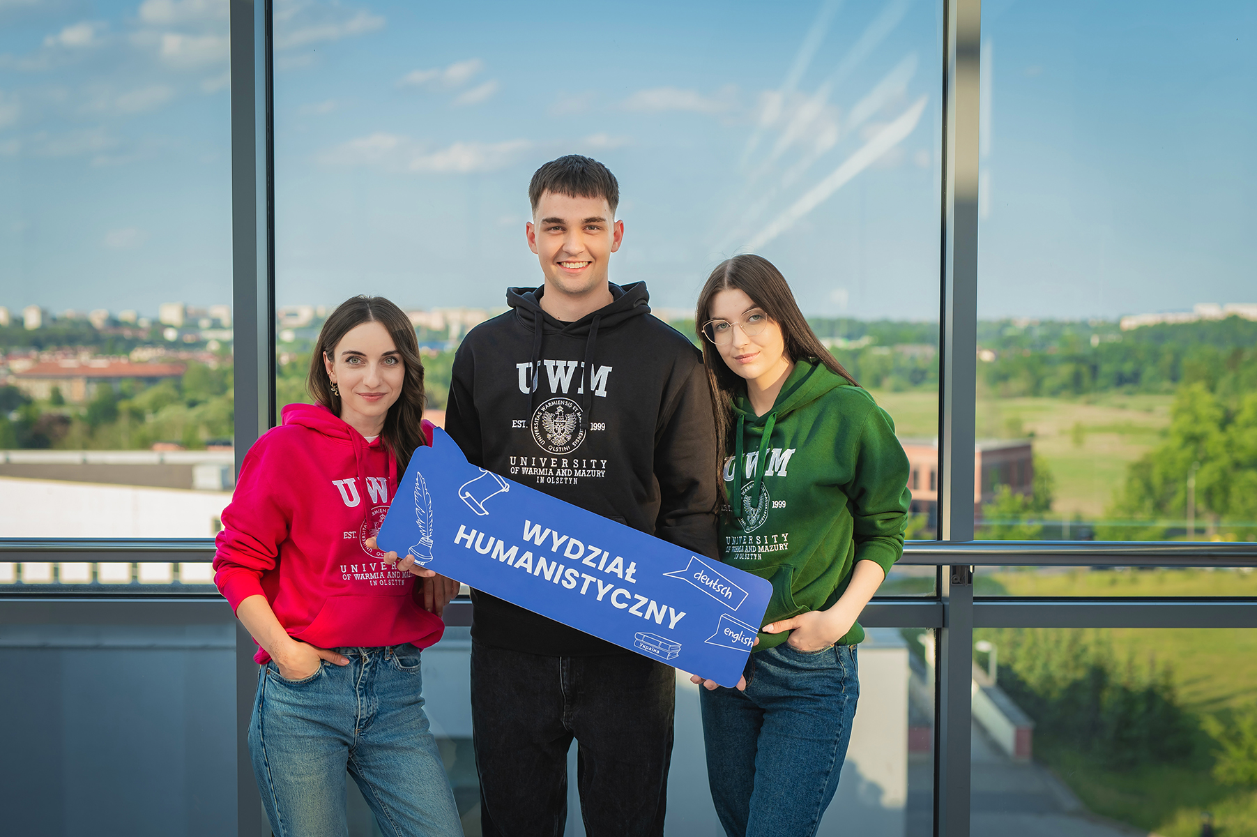 Troje studentów w kolorowych bluzach trzymających niebieską tabliczkę z napisem „Wydział Humanistyczny”