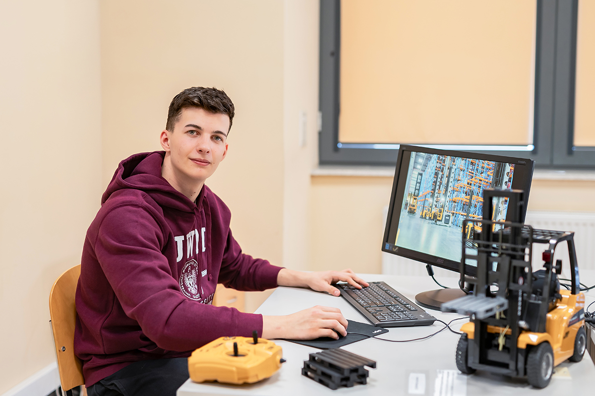 Osoba siedzi przy komputerze i obsługuje model wózka widłowego na stole