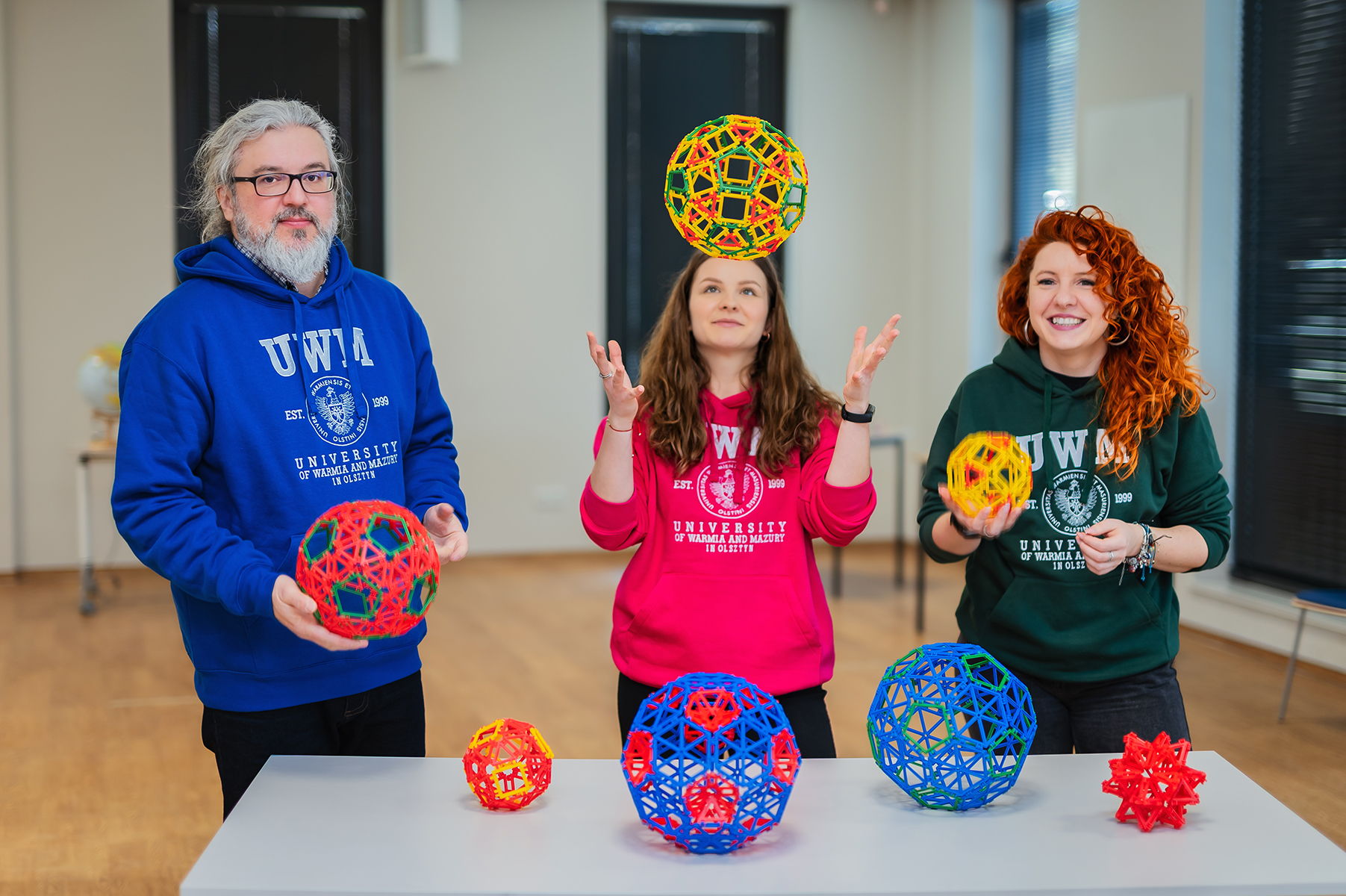 Trzy osoby prezentują kolorowe modele geometryczne w sali