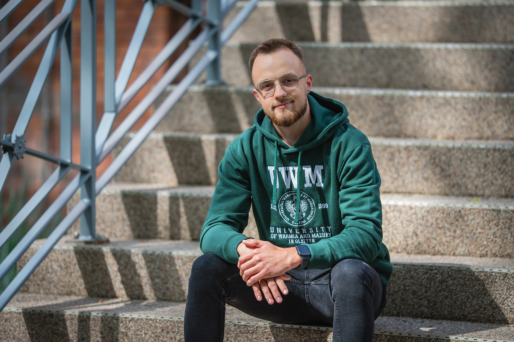 Osoba w zielonej bluzie z logo UWM siedzi na schodach przed budynkiem