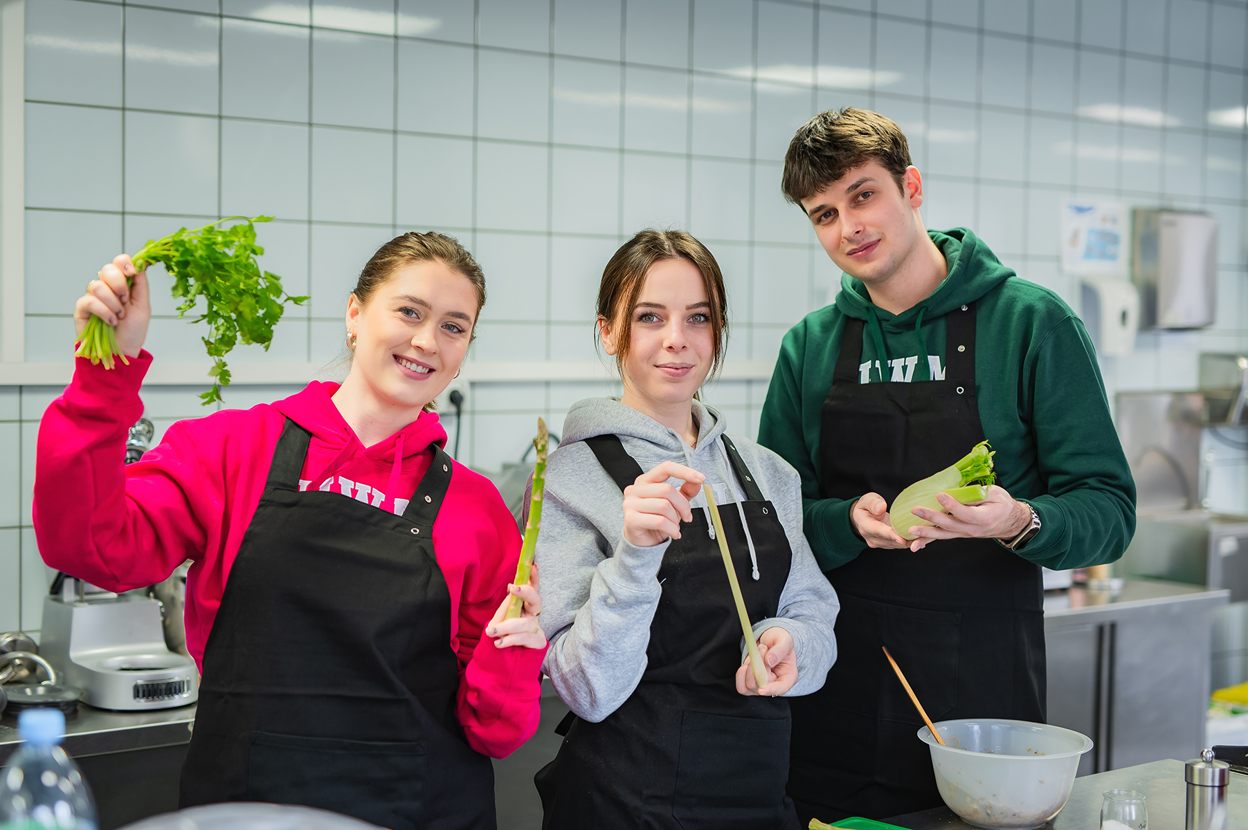 Trzech studentów gastronomii w fartuchach prezentuje warzywa w kuchni szkoleniowej