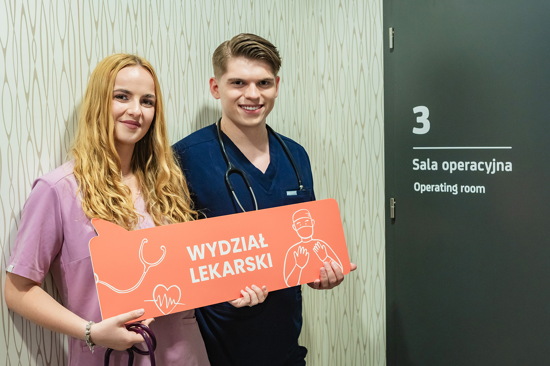 Dwoje studentów w strojach medycznych trzymających tabliczkę „Wydział Lekarski” przed salą operacyjną