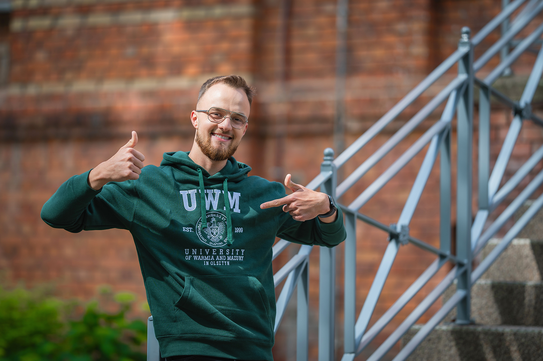 Osoba w zielonej bluzie z logo uczelni pokazuje gest aprobaty i wskazuje na napis UWM
