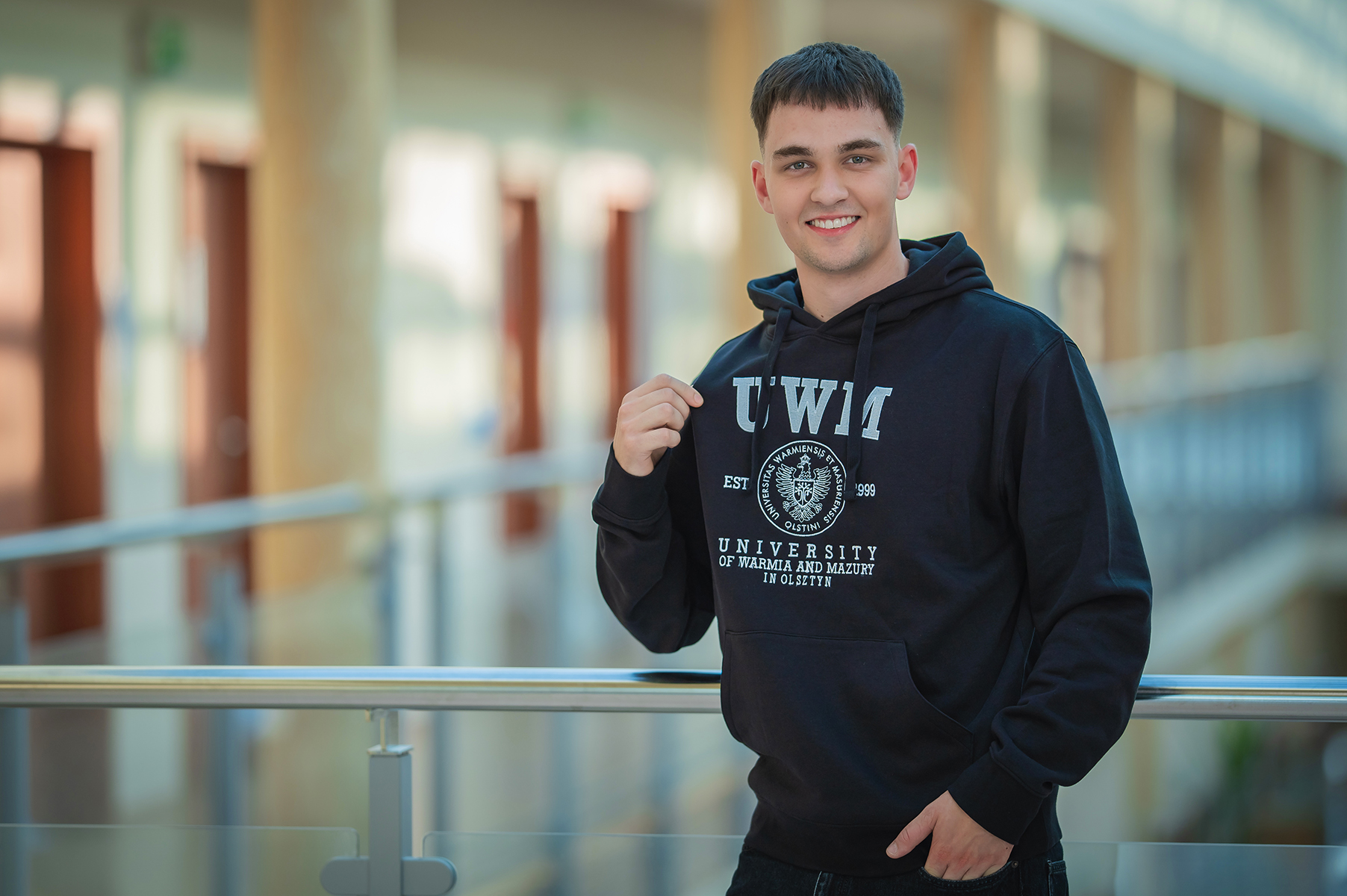 Uśmiechnięty student w czarnej bluzie z logo UWM na tle nowoczesnego korytarza