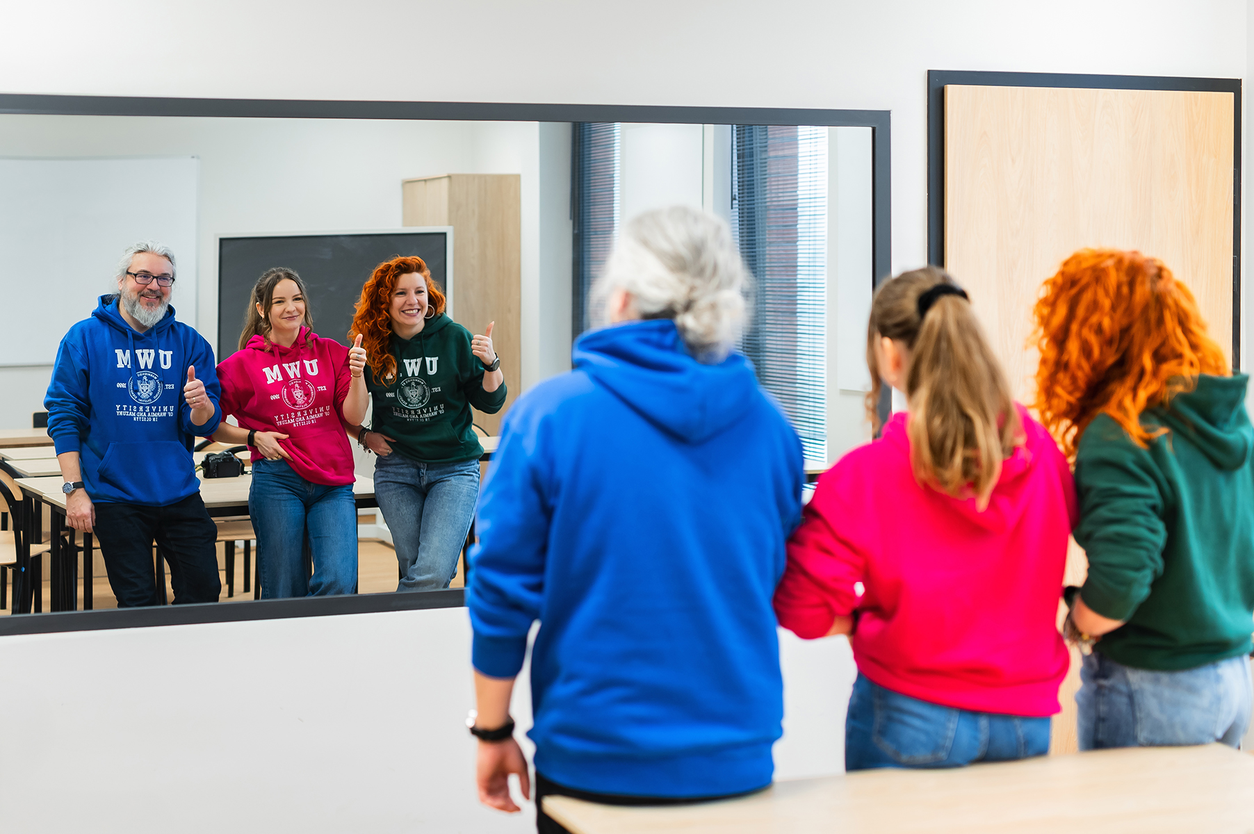 Grupa osób w bluzach z logo uczelni stoi w sali i patrzy w lustro