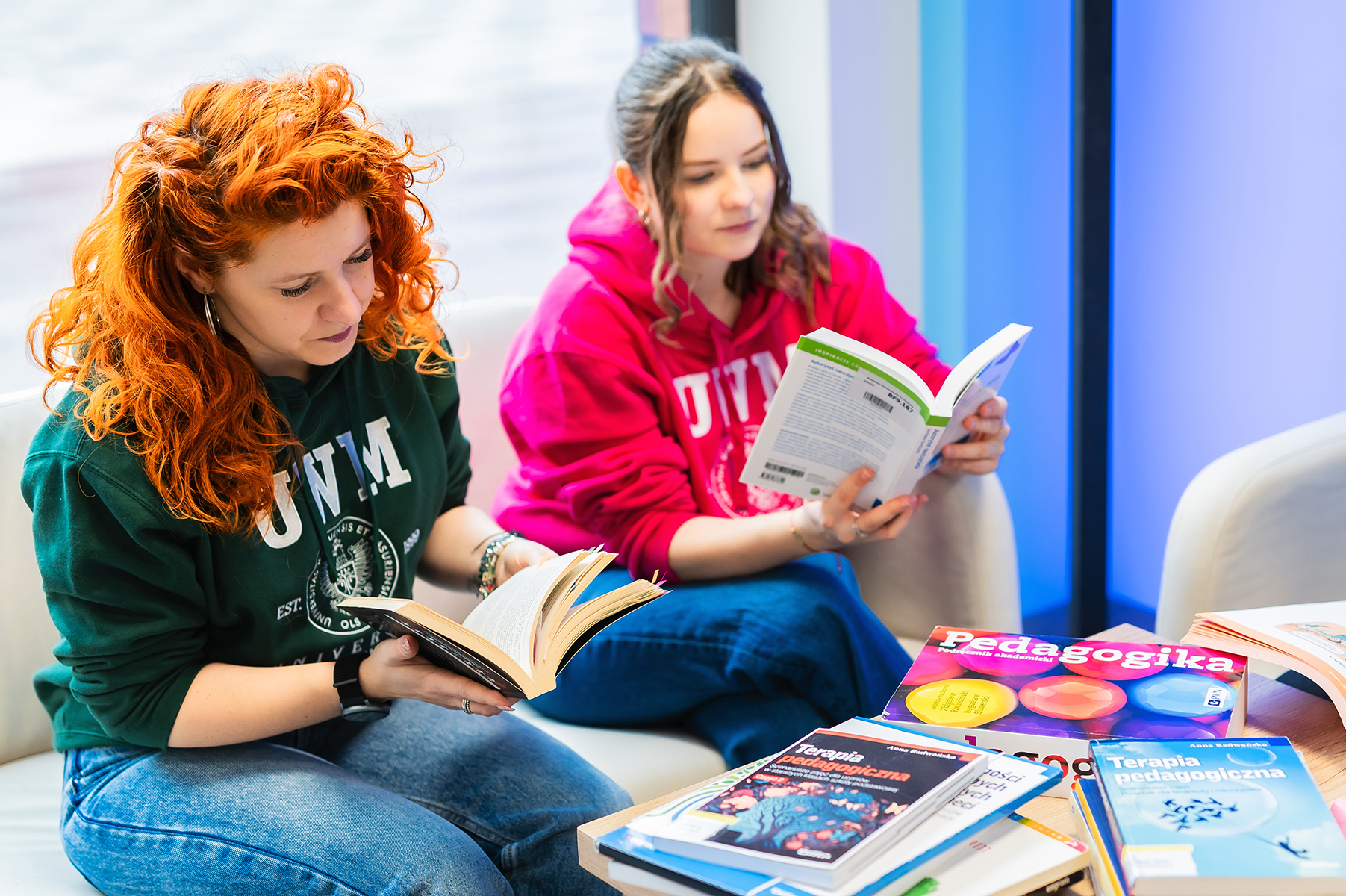 Dwie osoby siedzą na kanapie i czytają książki; na stole leżą podręczniki