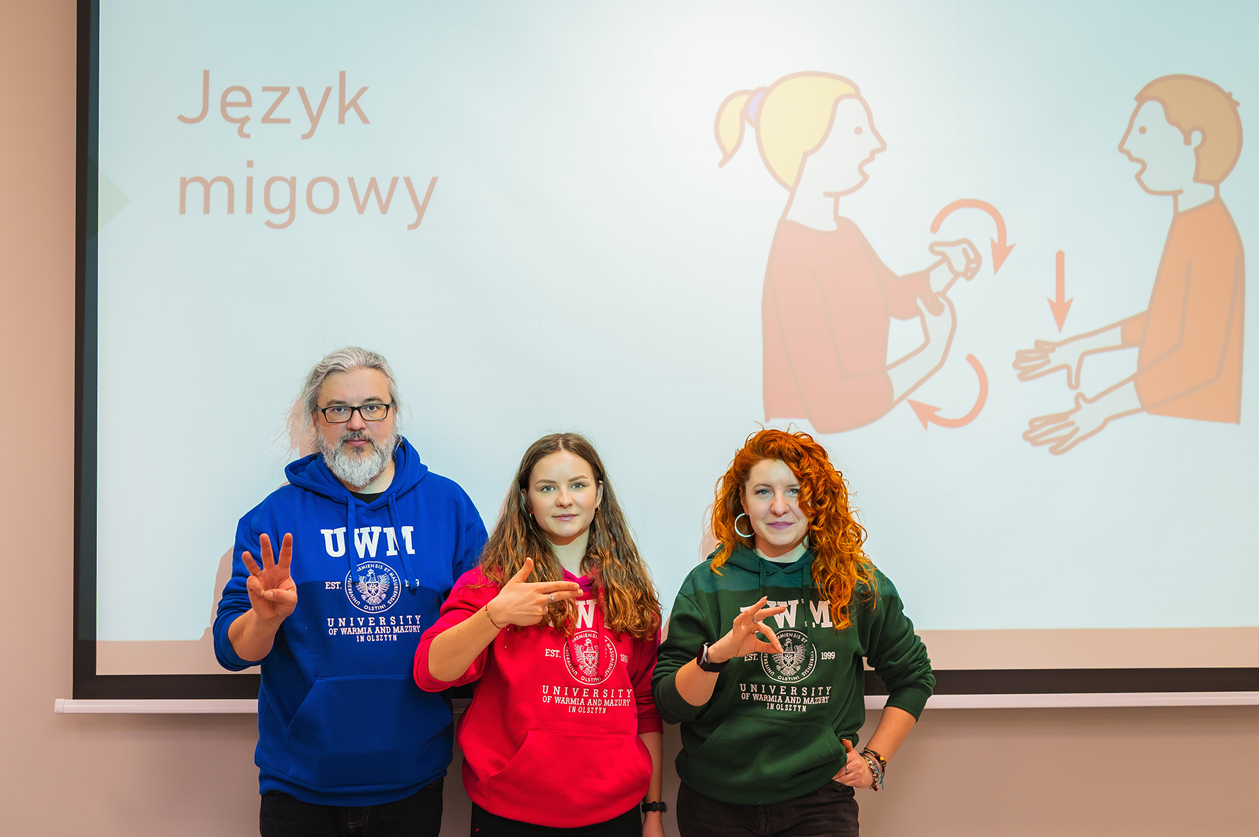 Trzy osoby stoją przed slajdem o języku migowym i pokazują znaki dłoni