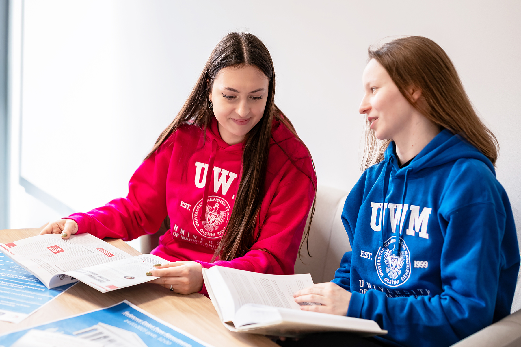 Dwie osoby w bluzach z logo UWM siedzą przy stole i wspólnie czytają materiały edukacyjne