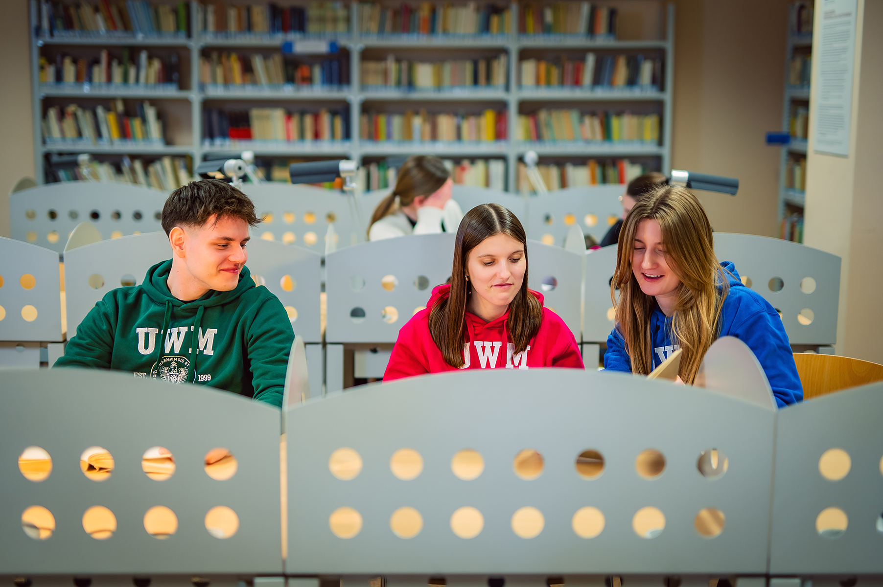 Studenci w bluzach UWM siedzą przy stanowiskach do nauki w bibliotece uniwersyteckiej