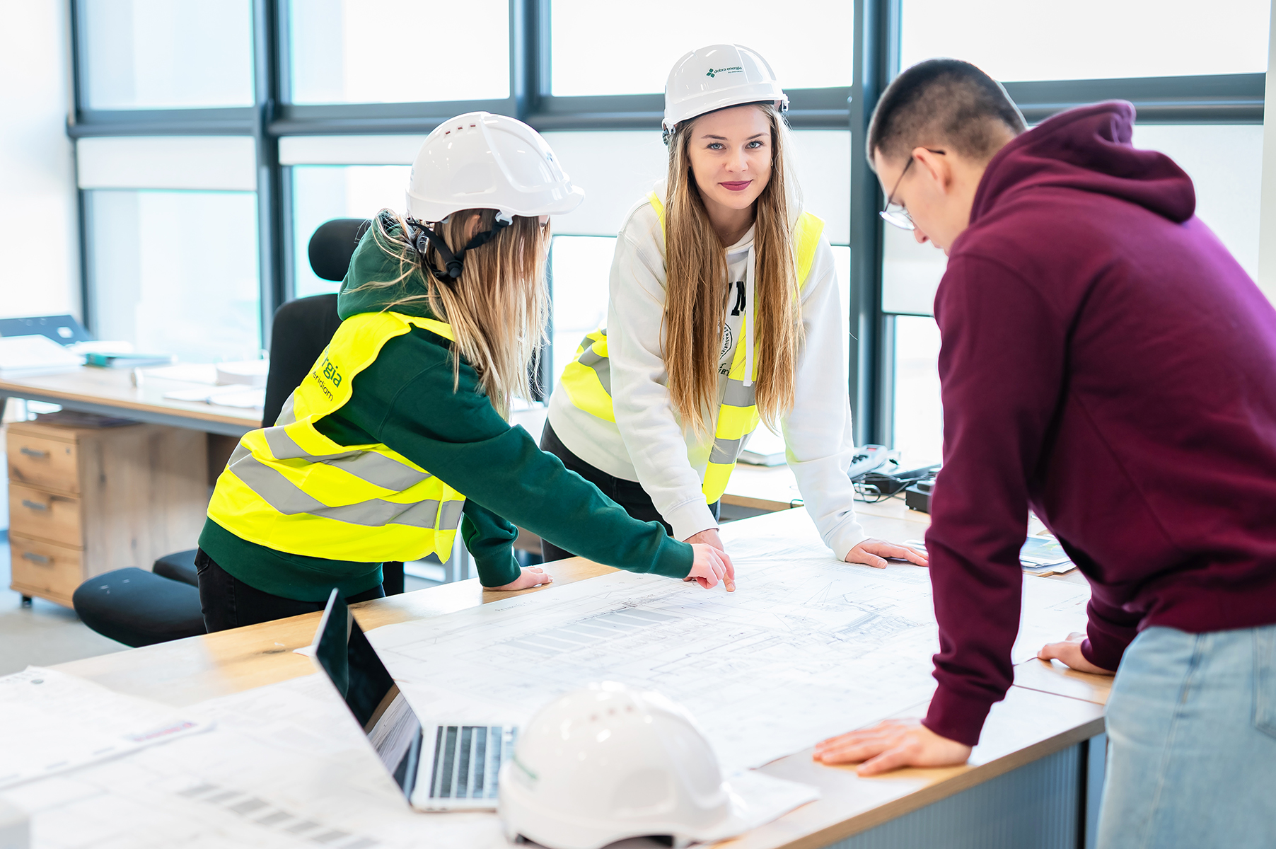 Osoby w kaskach i kamizelkach odblaskowych analizujące projekt techniczny na stole
