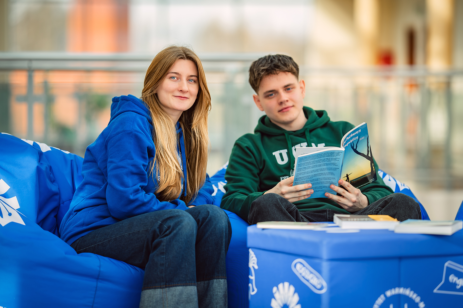 Studenci w bluzach UWM czytający książkę na pufach w strefie wypoczynku