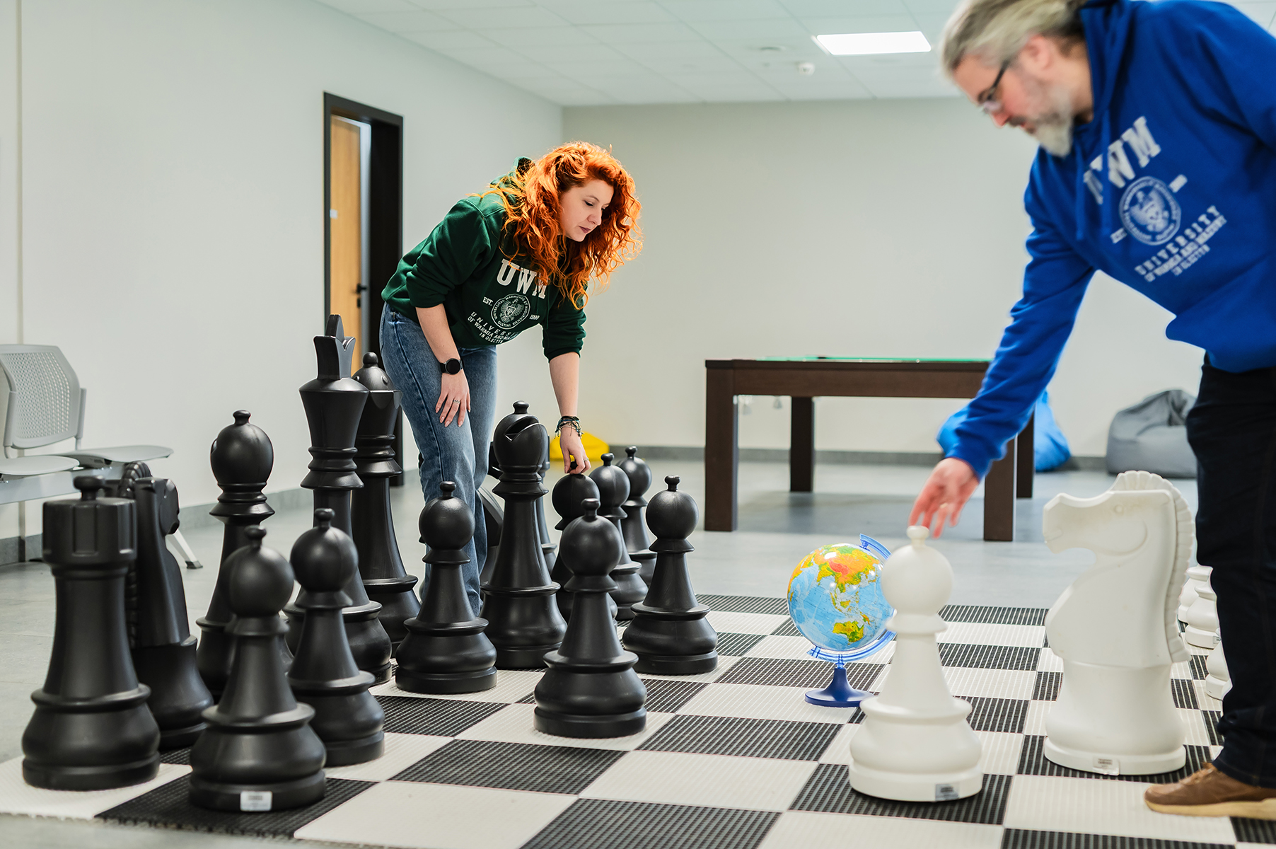 Dwie osoby ustawiają duże figury szachowe na planszy w sali