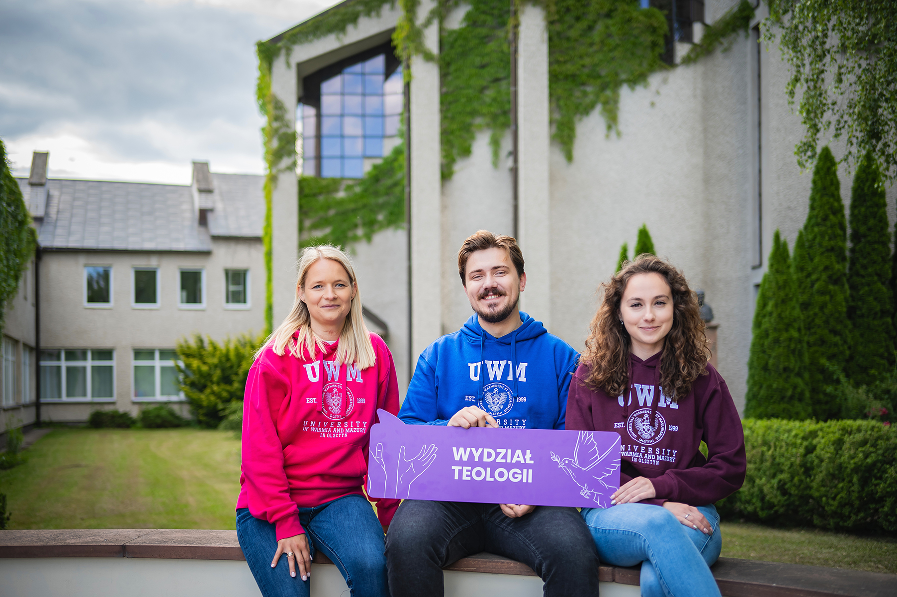 Trzy osoby siedzą na murku przed budynkiem i trzymają tablicę z napisem „Wydział Teologii”