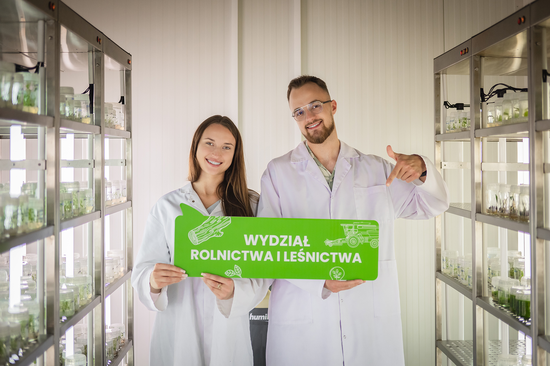 Dwie osoby stoją w laboratorium i trzymają zieloną tablicę z napisem "Wydział Rolnictwa i Leśnictwa"