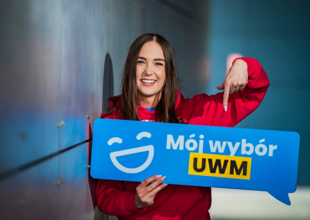 Dziewczyna trzyma w ręku tabliczkę z napisem "mój wybór UWM", z uśmiechem wskazuje palcem na trzymaną przez siebie tabliczkę.