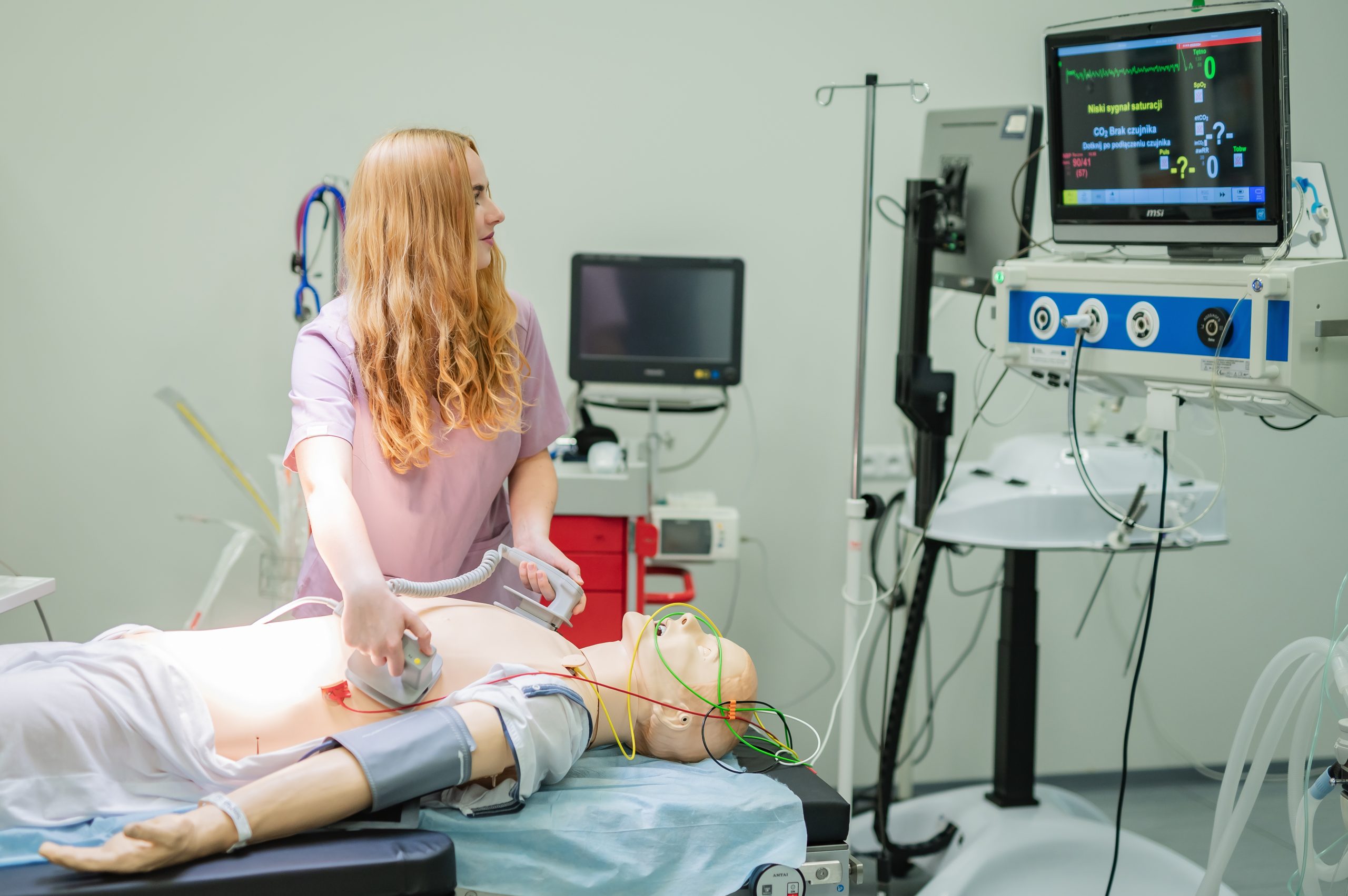 Kobieta w stroju medycznym przeprowadza symulację resuscytacji na manekinie leżącym na łóżku szpitalnym, używając defibrylatora. Manekin jest podłączony do przewodów i aparatury monitorującej parametry życiowe, a na monitorze obok wyświetlane są odczyty medyczne.