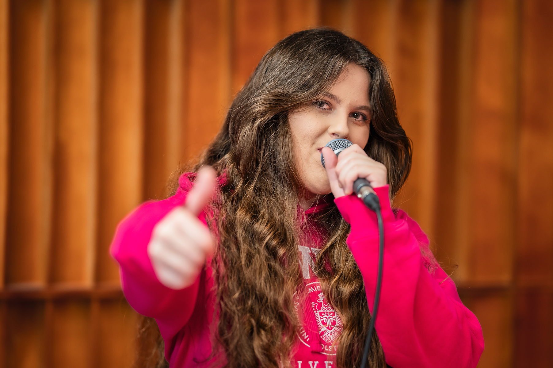 Osoba śpiewa do mikrofonu na scenie i pokazuje kciuk w górę