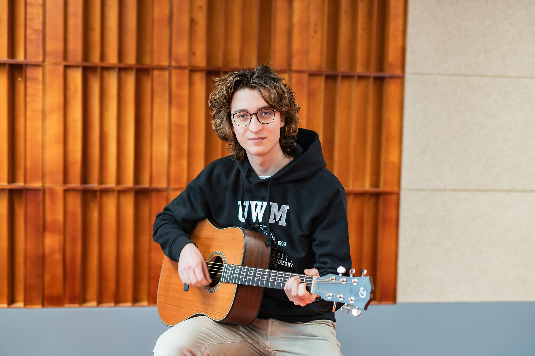 Osoba siedzi i gra na gitarze akustycznej w sali z drewnianą ścianą