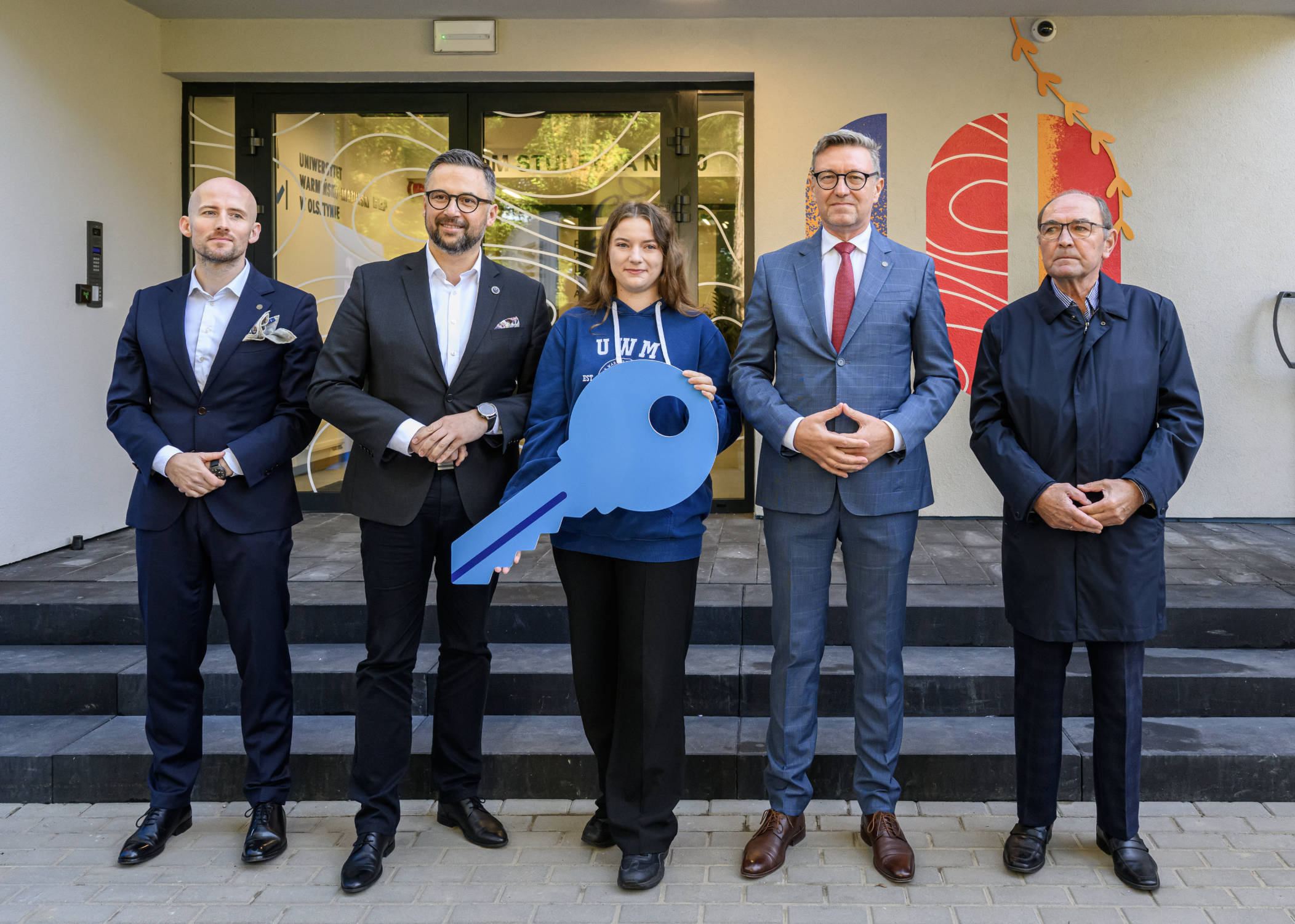 Otwarcie Domu Studenta nr 10 - przedstawiciele władz UWM, ministerstwa, studentów i wykonawcy budowy stoją przed nowo-otwartym budynkiem. Przedstawiciel samorządu studenckiego trzyma symboliczny klucz do akademika.