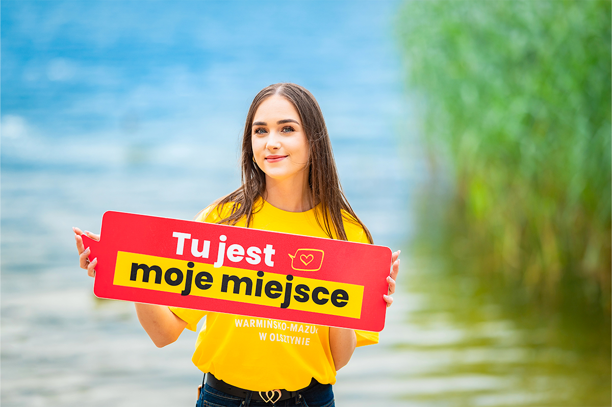 Młoda kobieta w żółtej koszulce nad jeziorem trzyma tabliczkę z napisem „Tu jest moje miejsce”