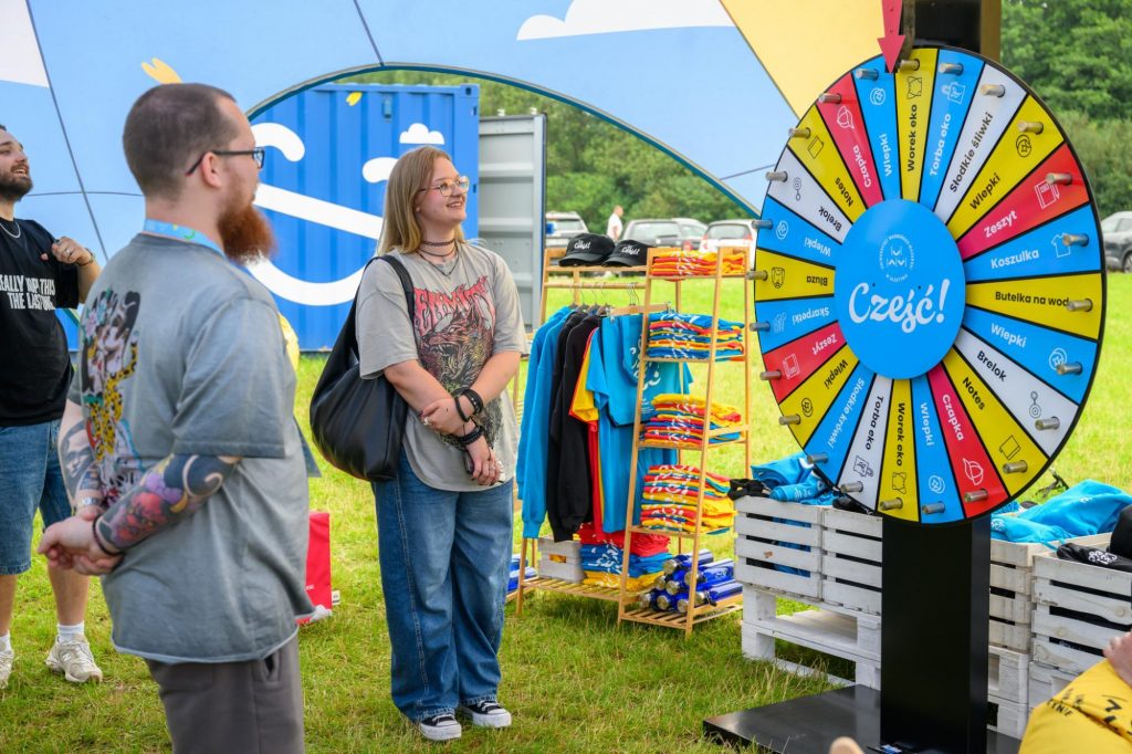 Studenci stoją przy kole fortuny z nagrodami promocyjnymi uczelni podczas wydarzenia plenerowego.